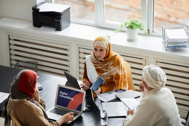 Three businesswomen in hijabs discussing digital advertising at an office table. Direct publisher buying versus platform buying: commitments